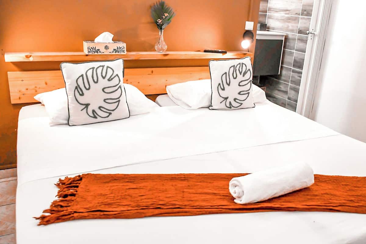 A neatly made bed with white bedding and two decorative pillows, a wooden headboard, a bedside table with a tissue box, and a view of a tiled wall and window.