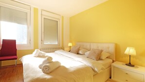 A hotel room with a bed, a nightstand, a lamp, a chair, and two windows with blinds. - Apartment Bella Art (Belgrade)