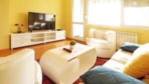 A modern living room with a flat-screen TV, white sofas, a coffee table, and a window with blinds. - Apartment Bella Art (Belgrade)