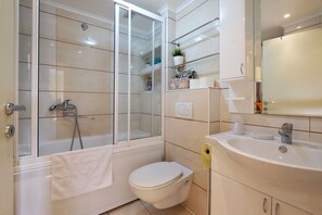 A modern bathroom with a glass-enclosed shower, a white toilet, a sink with a mirror, and a shelf with toiletries. - Apartment Bella Art (Belgrade)