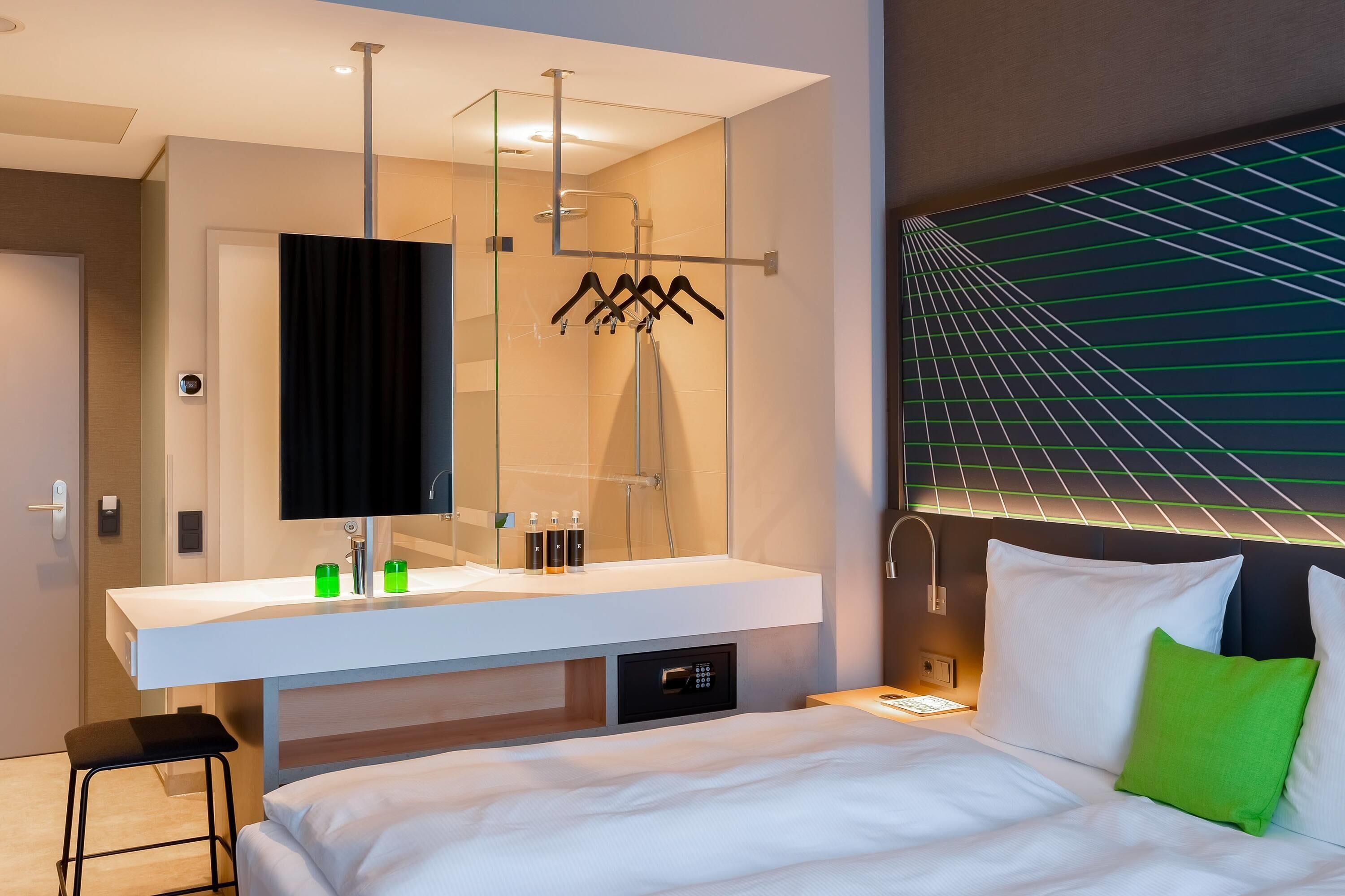 Ett modernt hotellrum med en stor säng, en vit sminkbord med spegel, en dusch och ett sängbord med en lampa.