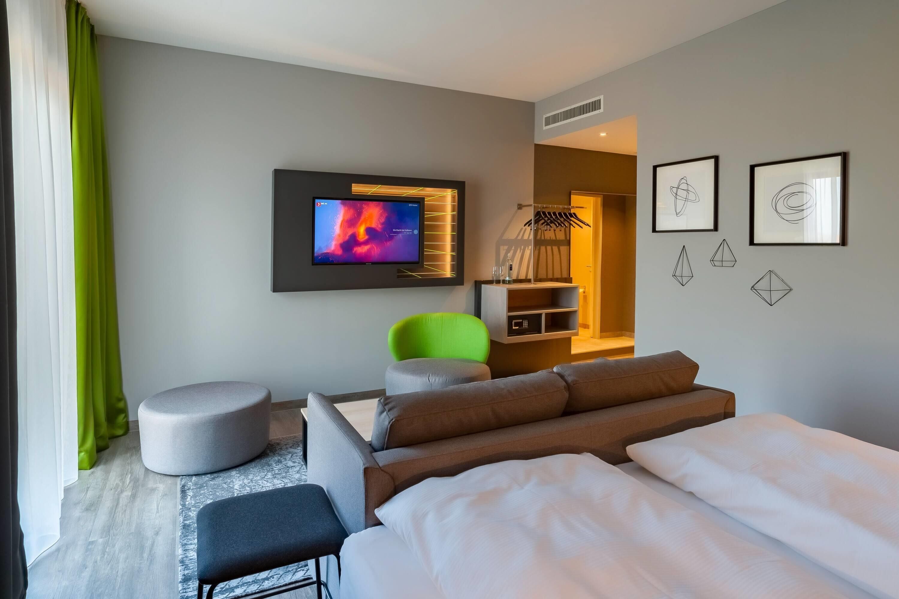 A modern hotel room with a flat-screen TV, a grey sofa, a green chair, and a bed with white bedding.