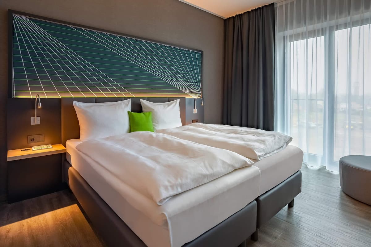 Een moderne hotelkamer met een groot bed, een hoofdeinde met een geometrisch lichtontwerp, een nachtkastje met een lamp, en uitzicht op het omliggende landschap door dunne gordijnen.