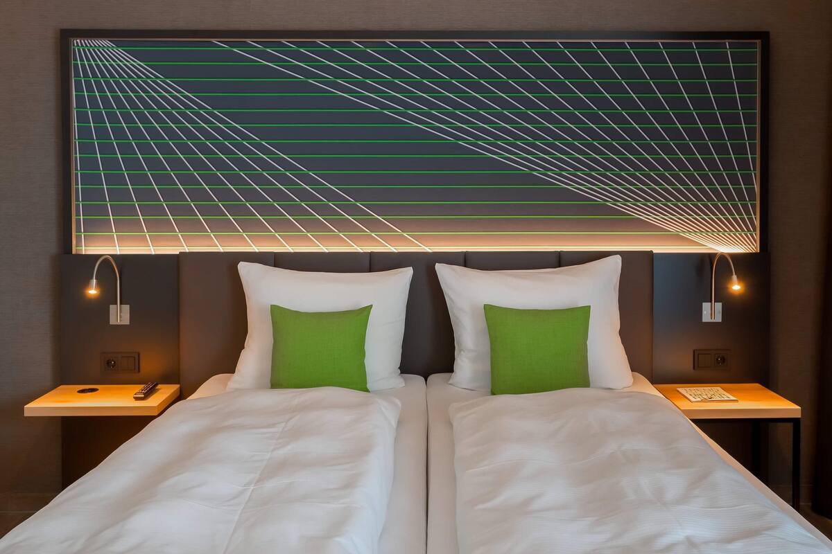 Een tweepersoonsbed met witte beddengoed en groene kussens, met nachtkastjes met lampen ernaast, in een kamer met een geometrische lichtinstallatie aan de muur.