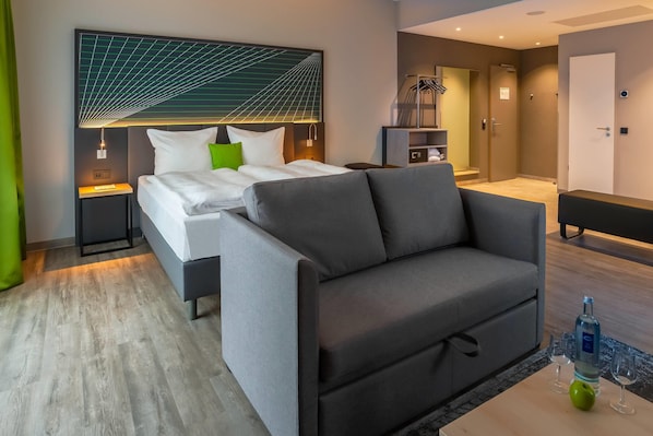 Une chambre d’hôtel moderne avec un lit, un canapé gris, une petite table avec une bouteille et des verres, et une armoire.