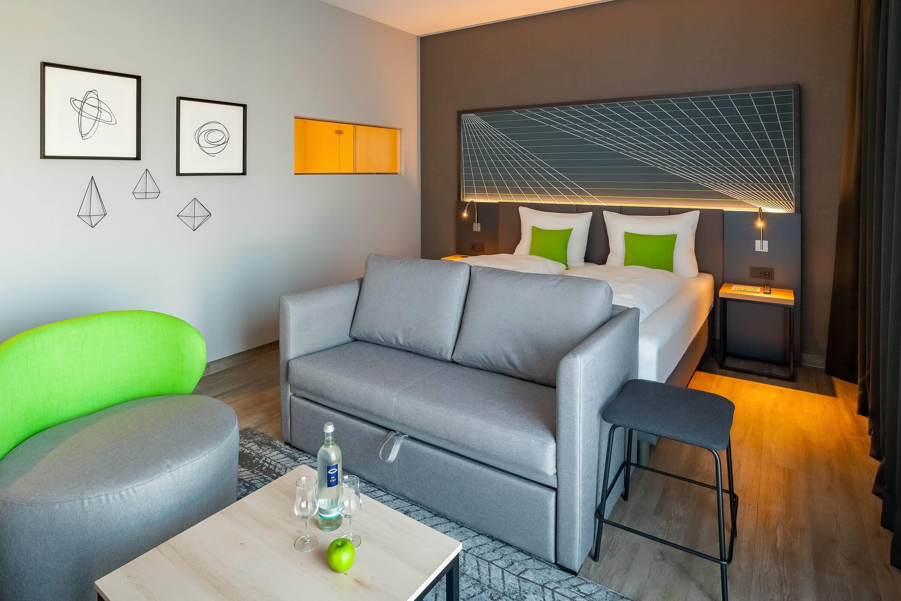 Ein modernes Hotelzimmer mit grauem Sofa, einem grünen Sessel, einem Holztisch mit einer Flasche und Gläsern sowie einem Bett mit einem einzigartigen Kopfteildesign.
