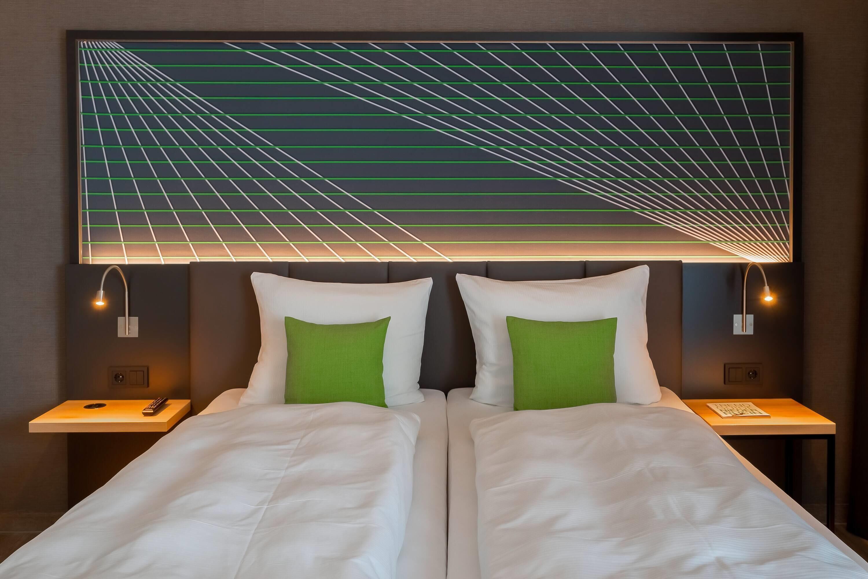 A double bed with white bedding and green pillows, flanked by nightstands with lamps, against a wall with a geometric light fixture.