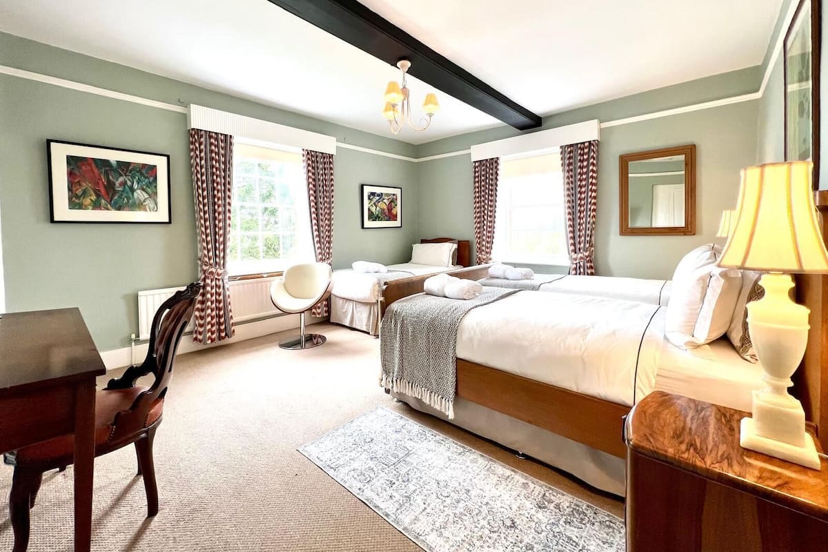 A hotel room with a bed, a desk, a chair, a lamp, and two windows with curtains.