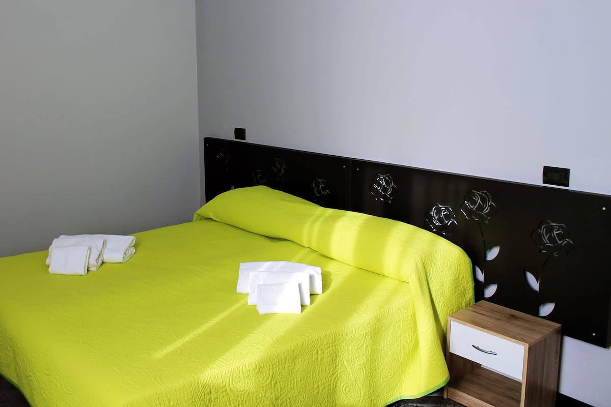 A neatly made bed with a bright green bedspread and white towels, a wooden nightstand with a drawer, and a wall with a decorative metal design.