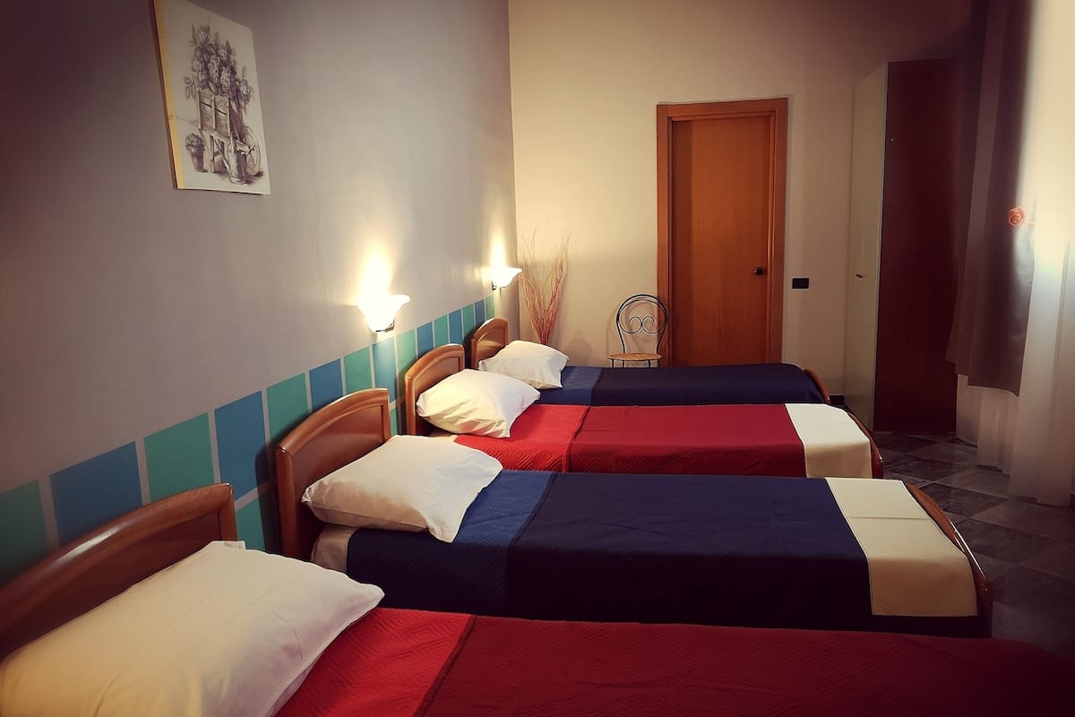 A hotel room with three single beds, each with a different colored bedspread. There is a wooden door and a wall-mounted light fixture.