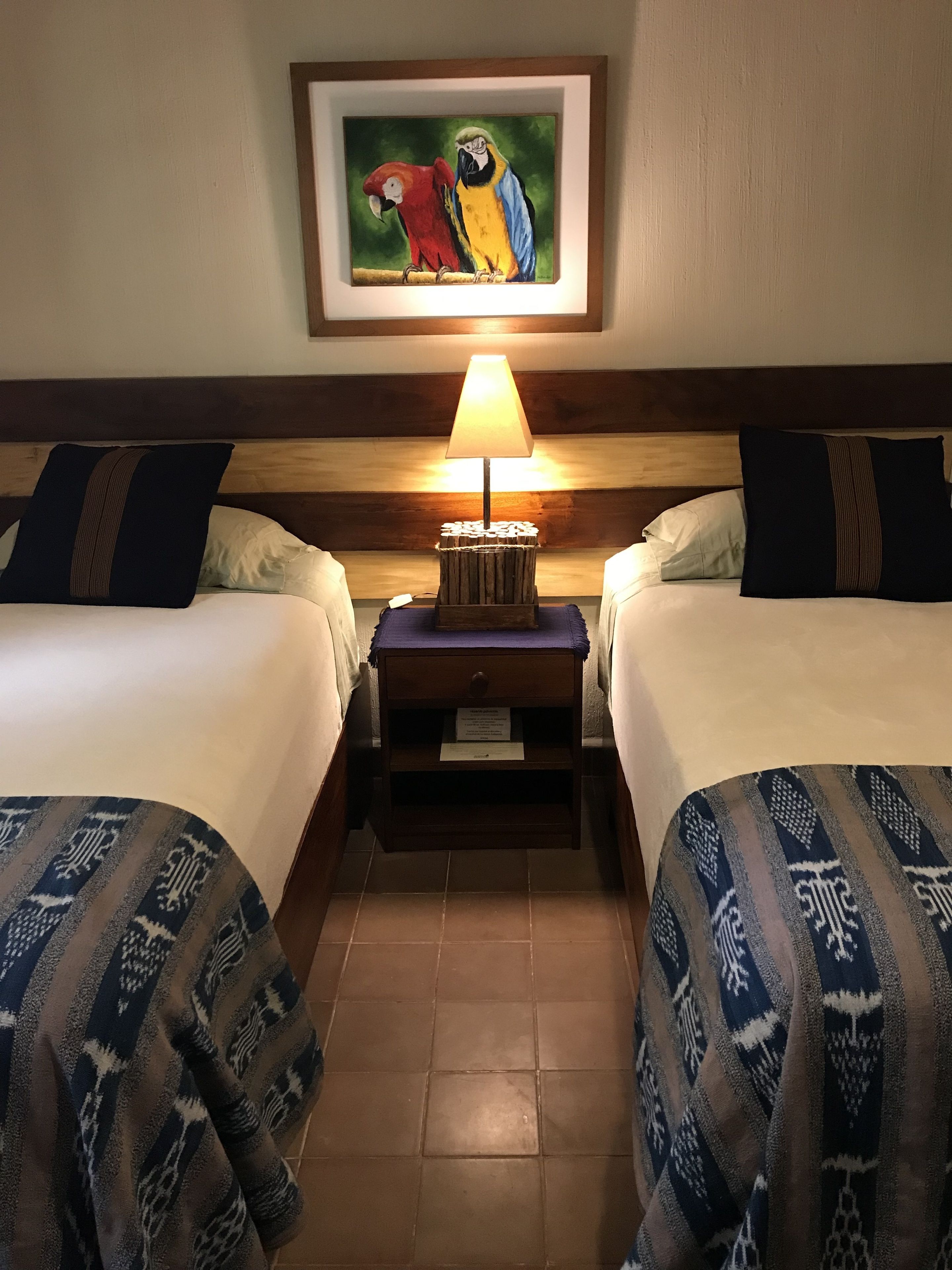 A hotel room with two beds, a nightstand with a lamp, a framed painting of parrots, and patterned bedspreads.