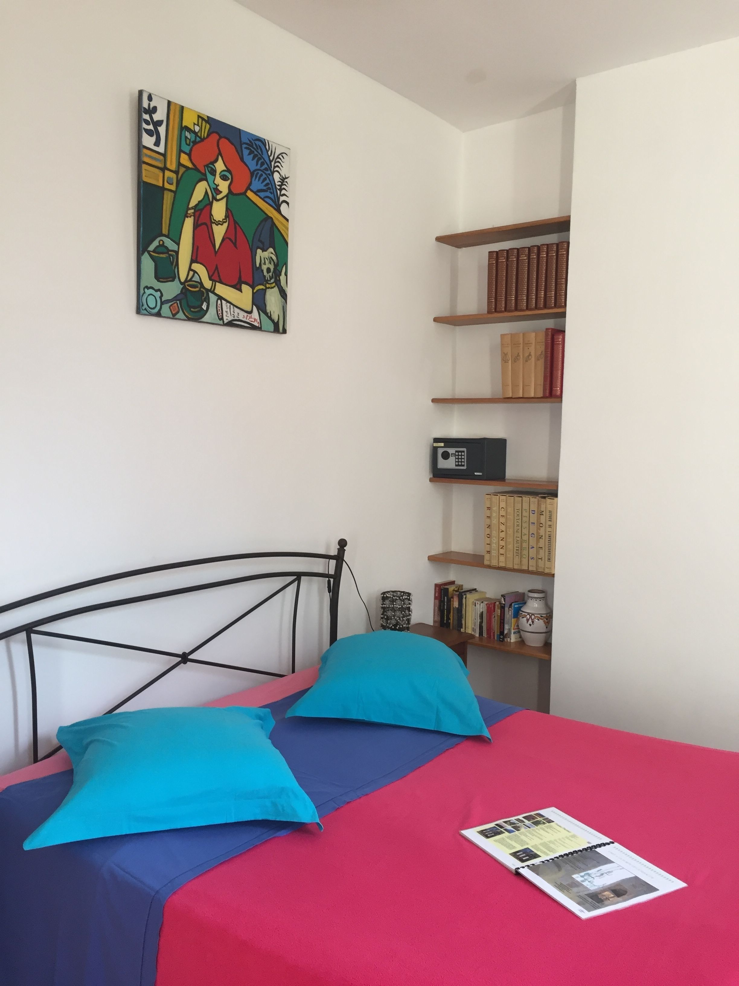 A bedroom with a bed, two pillows, a bookshelf, and a painting on the wall.