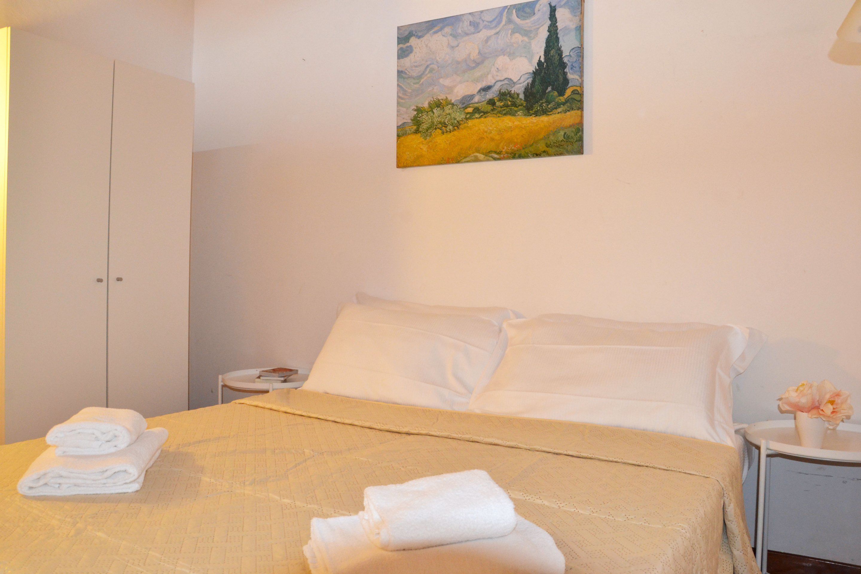 A neatly made bed with white linens and towels, a painting on the wall, and a small bedside table with a vase of flowers.