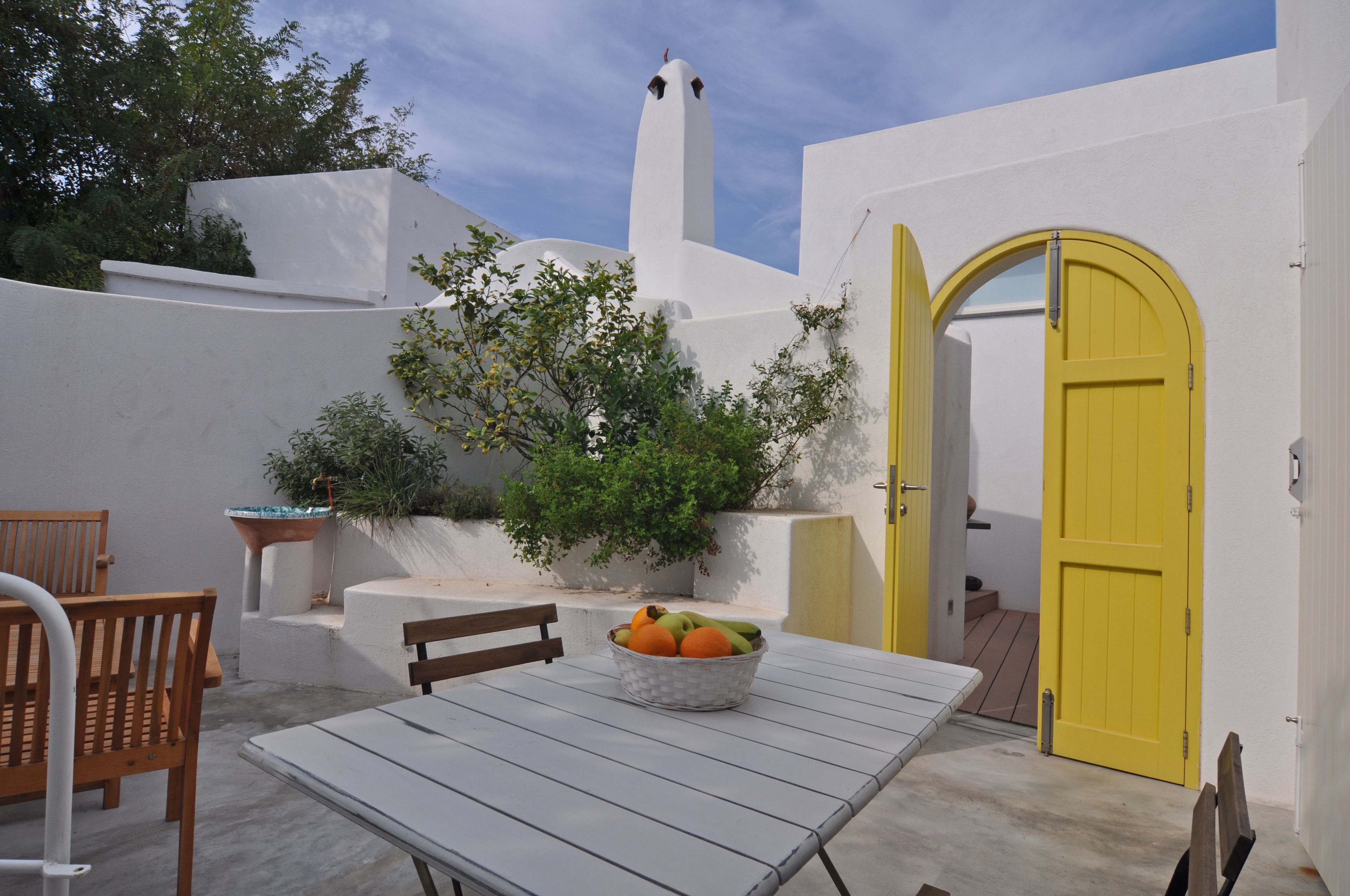 A white-walled building with a yellow door, a table with a fruit bowl, and a bench.