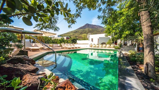 A clear pool with a mountain in the background, surrounded by lounge chairs and rocks. - Il Gabbiano Relais (Lipari)