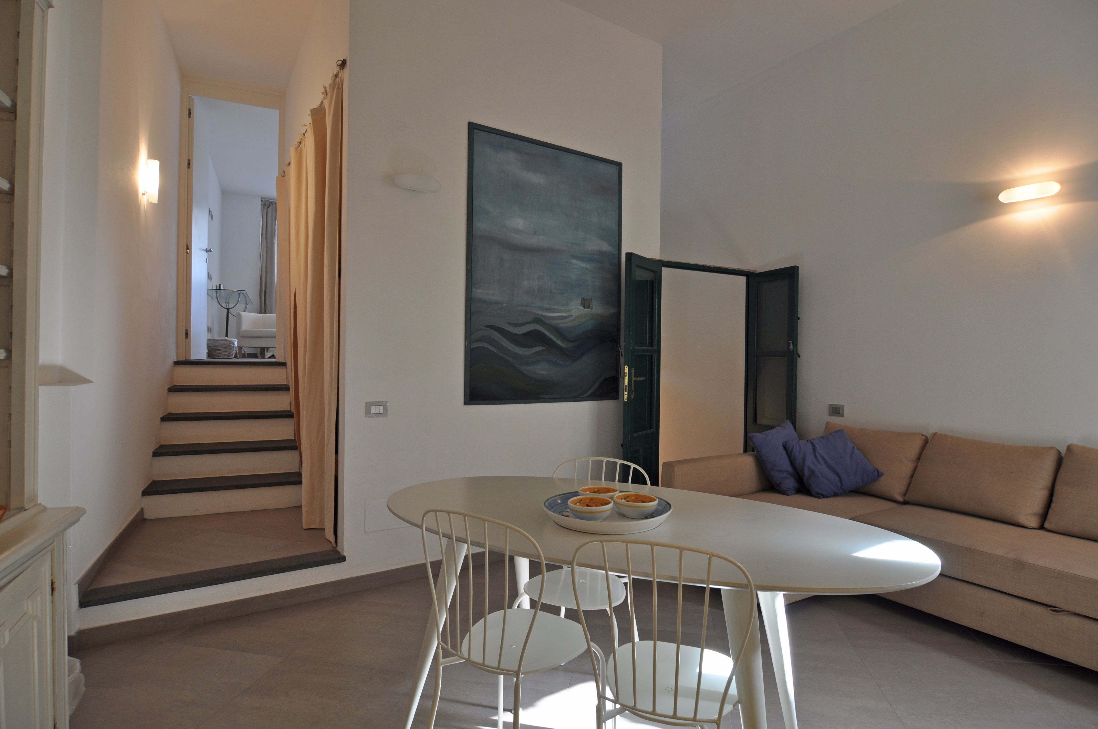 A modern living room with a white sofa, a dining table with chairs, and a staircase leading to another room.