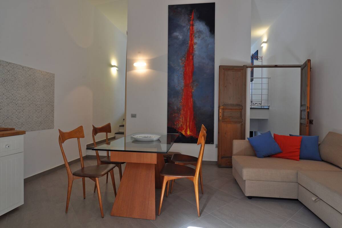 A modern living room with a glass dining table, wooden chairs, a beige sofa with blue and red cushions, and a large abstract painting on the wall.
