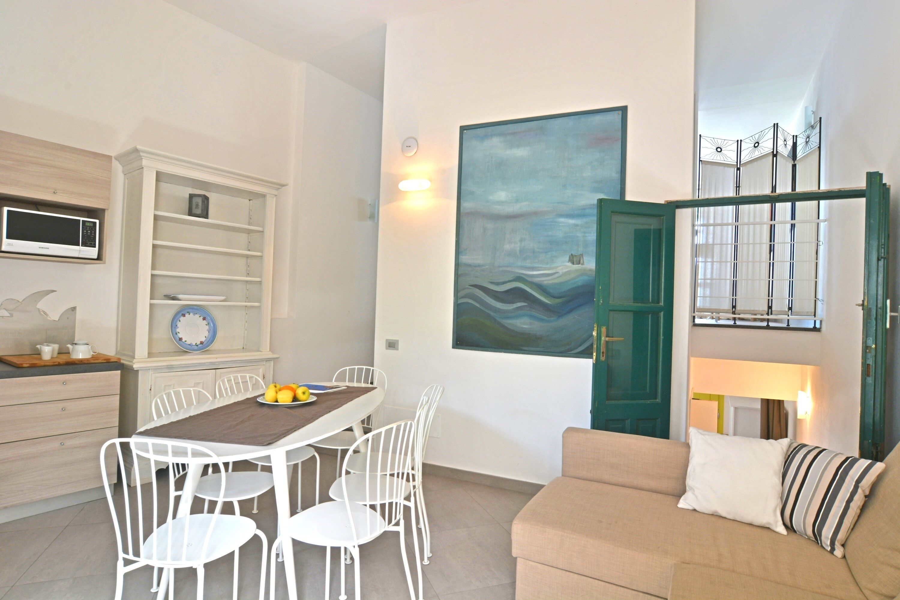 Una sala moderna con un sofá color beige, una mesa de comedor con sillas blancas, una zona de cocina con microondas y un cuadro abstracto grande en la pared.