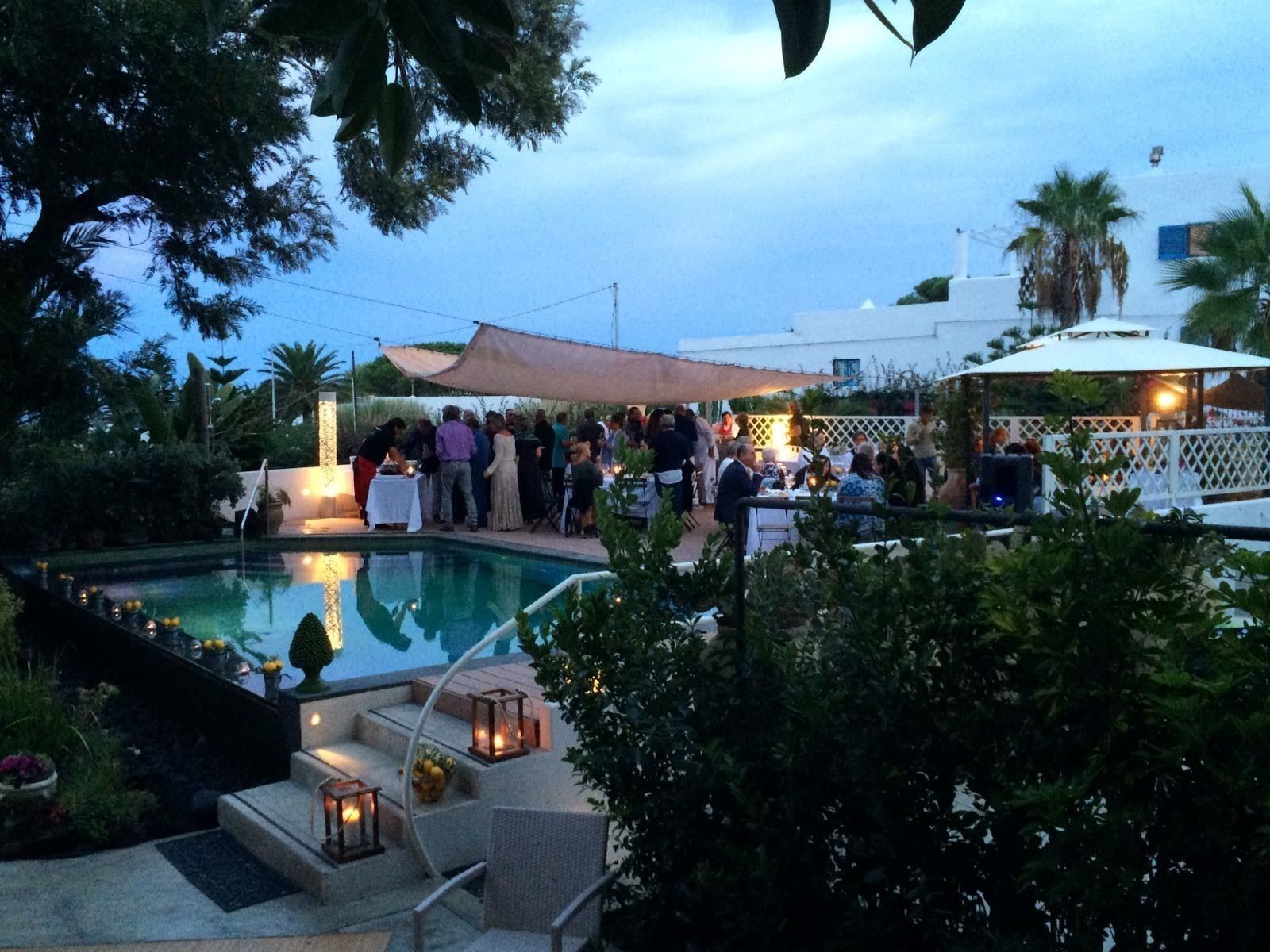 An outdoor pool area with a gathering of people, a covered seating area, and a white building in the background.