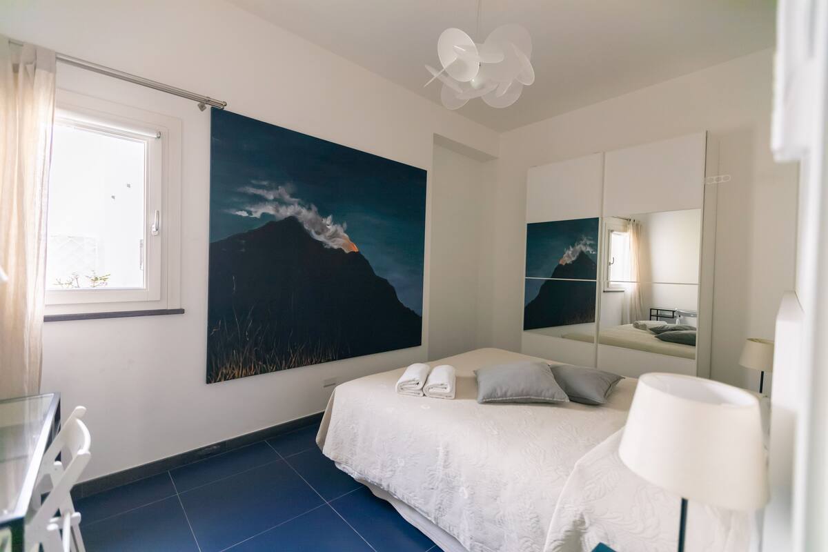A hotel room with a bed, a large painting of a mountain, a window with curtains, a mirror, and a white lamp.