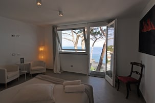 Una habitación de hotel moderna con una cama grande, una zona de estar con sillas, una mesita y vista al mar a través de una puerta abierta.
