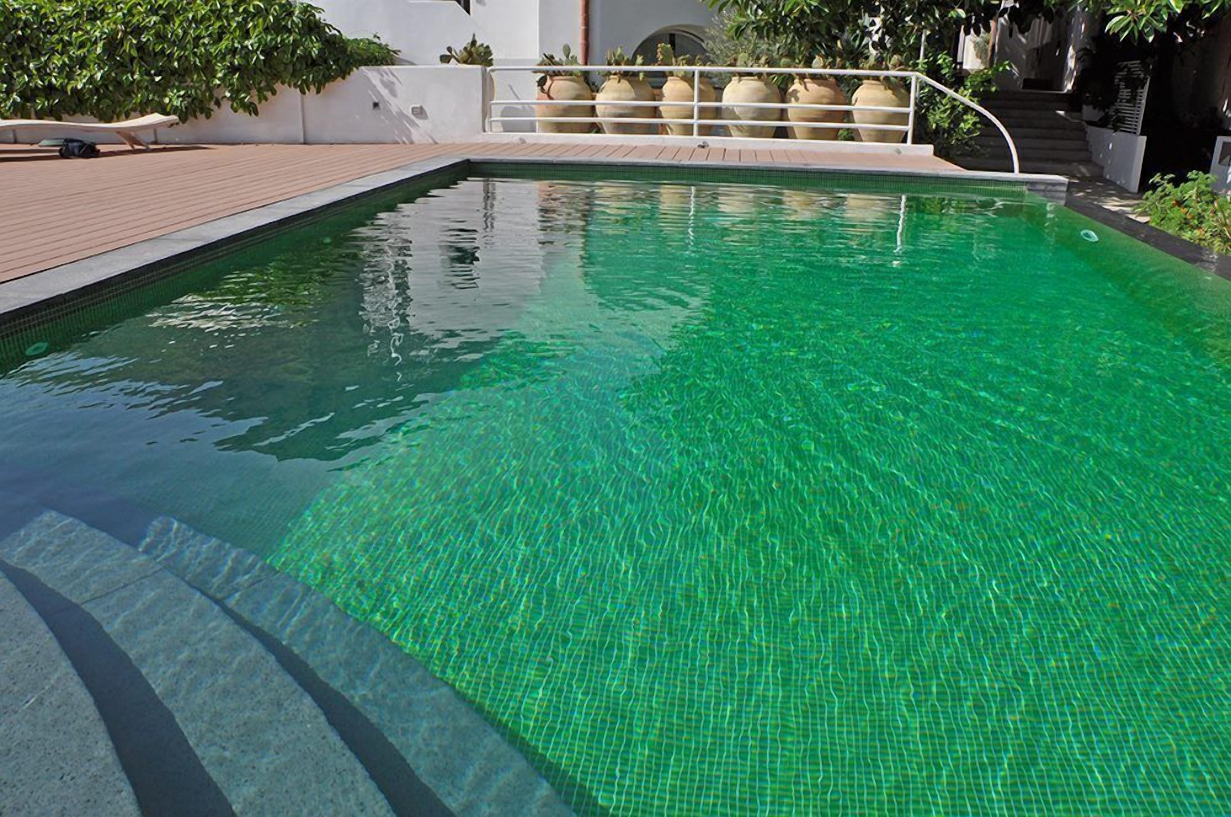 A rectangular swimming pool with clear water, surrounded by a tiled deck and a white wall with a metal railing.