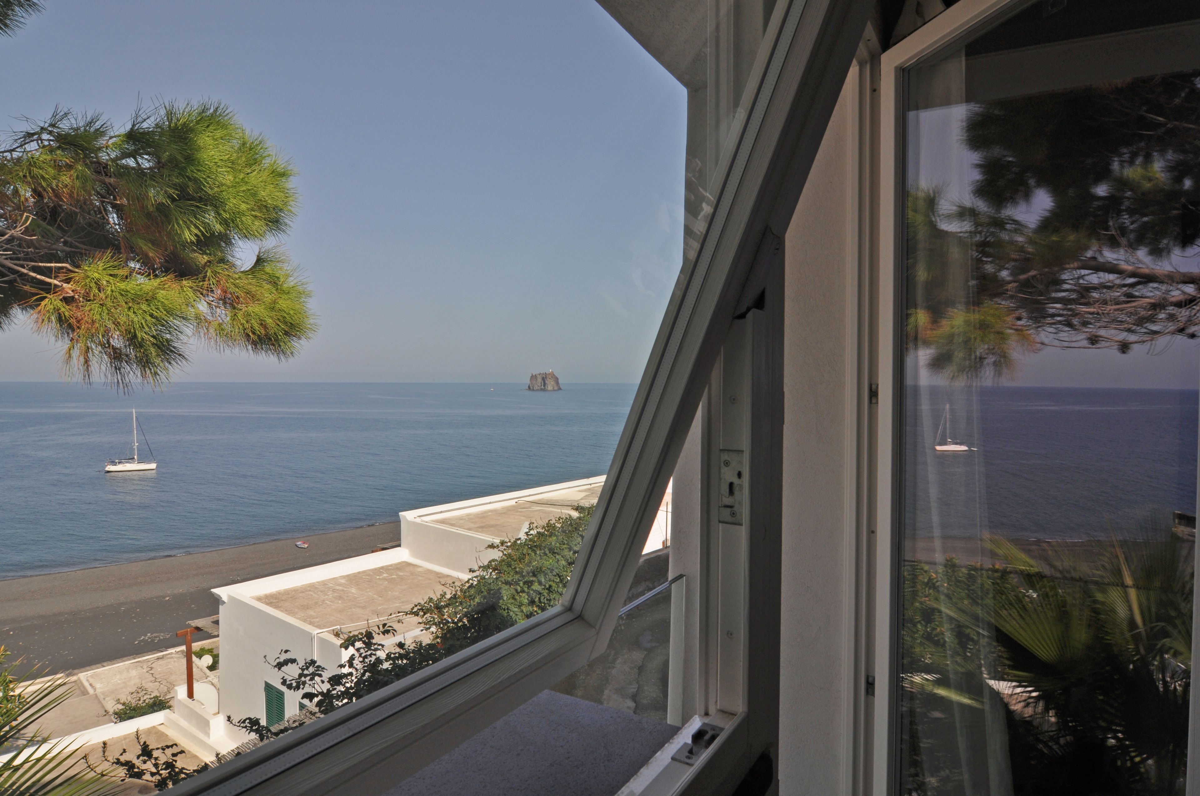 Vista costera desde una ventana que da a una playa con un velero y una isla distante.
