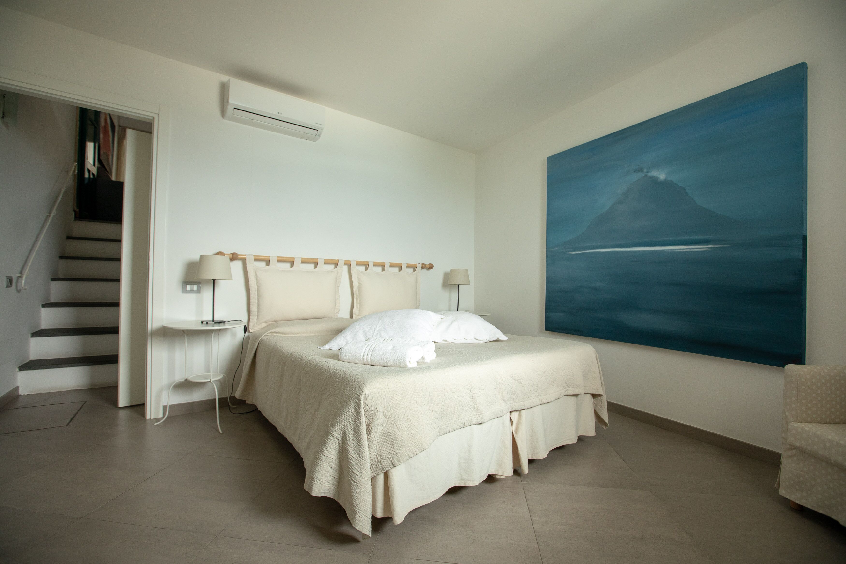 A bedroom with a bed, nightstands, a painting, a chair, and a staircase.
