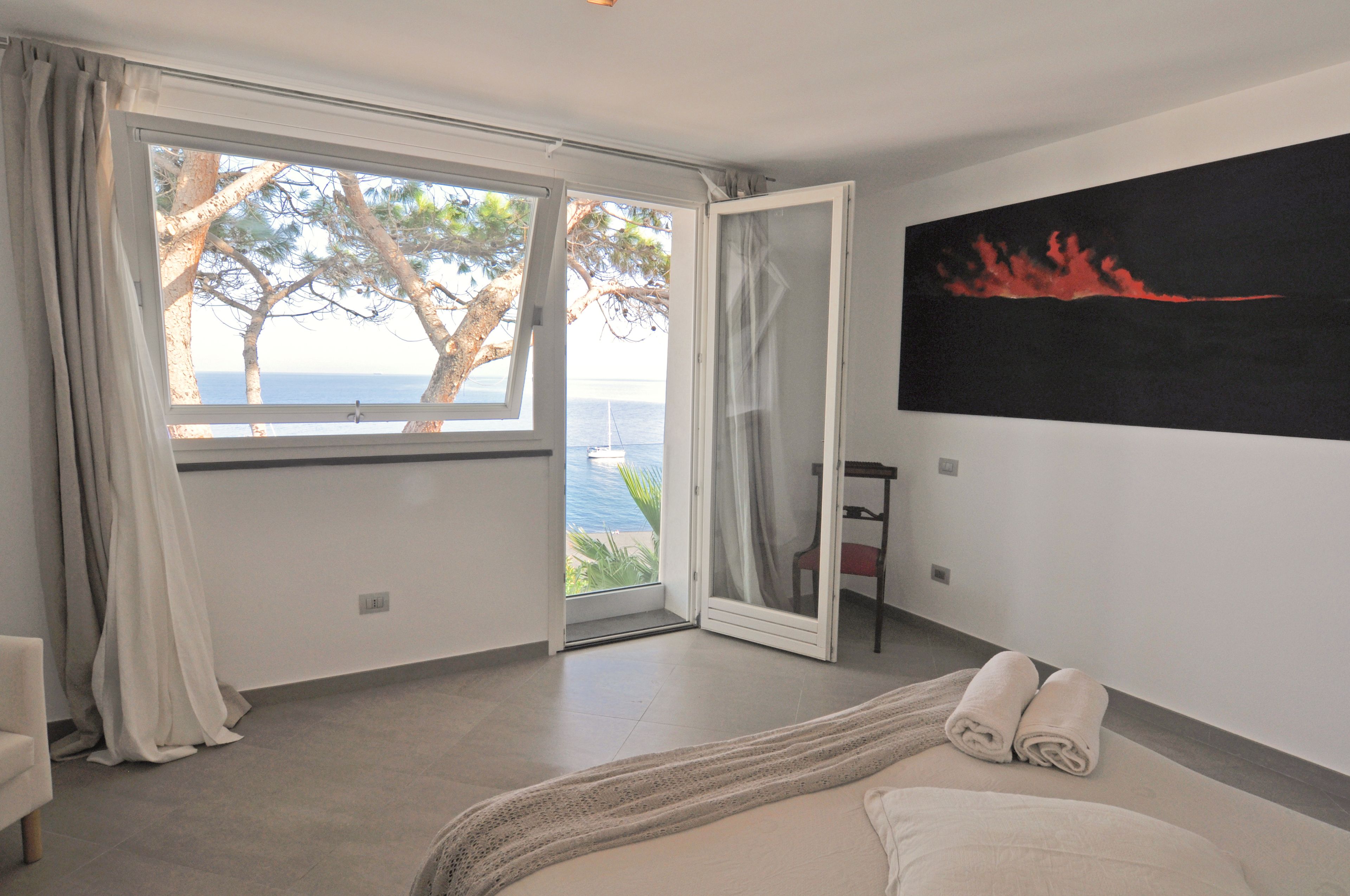 A bedroom with a large window, a bed, a chair, and a painting.