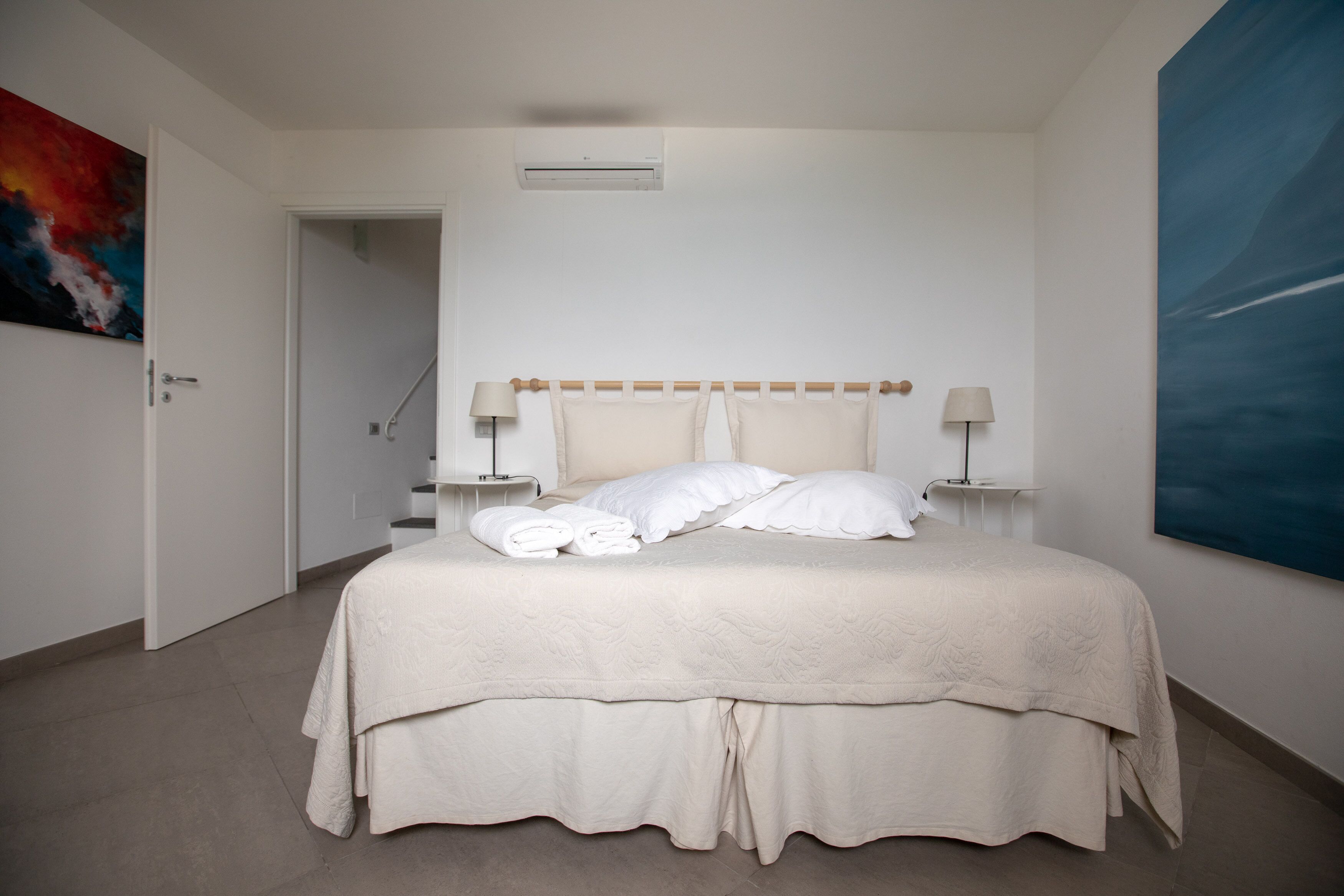 A neatly made bed with white linens, a headboard, bedside lamps, and a large abstract painting on the wall.