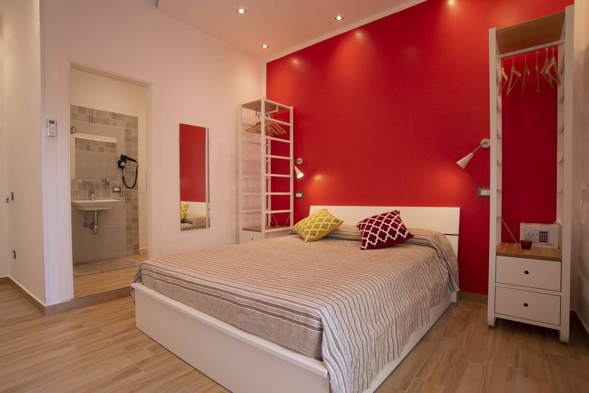 A bedroom with a bed, a wardrobe, a bedside table, a lamp, and a bathroom with a shower and sink.