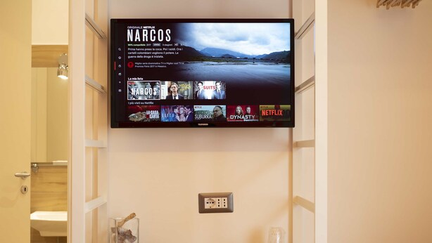 A hotel room with a mounted TV displaying a Netflix interface, a wooden desk with a glass container, and a wall socket. - Vanvitelli Domus (Naples)