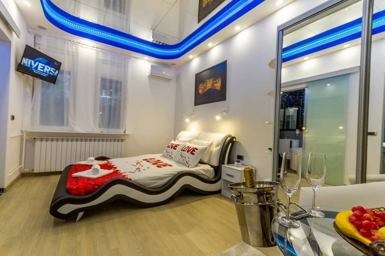 A modern hotel room with a bed featuring a wave design, a minibar, a dining table with a fruit bowl, and a TV displaying 'NIVERS'.