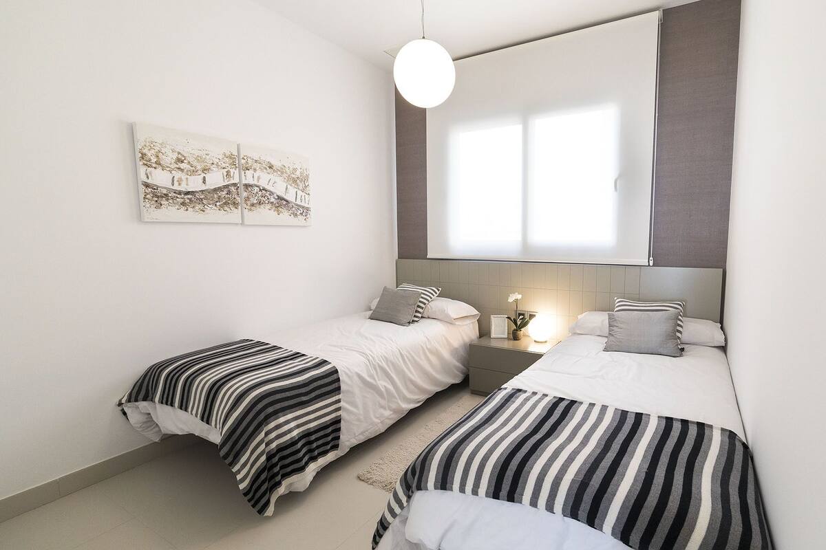 A bedroom with two beds, a nightstand, a wall-mounted light, and a window with blinds.