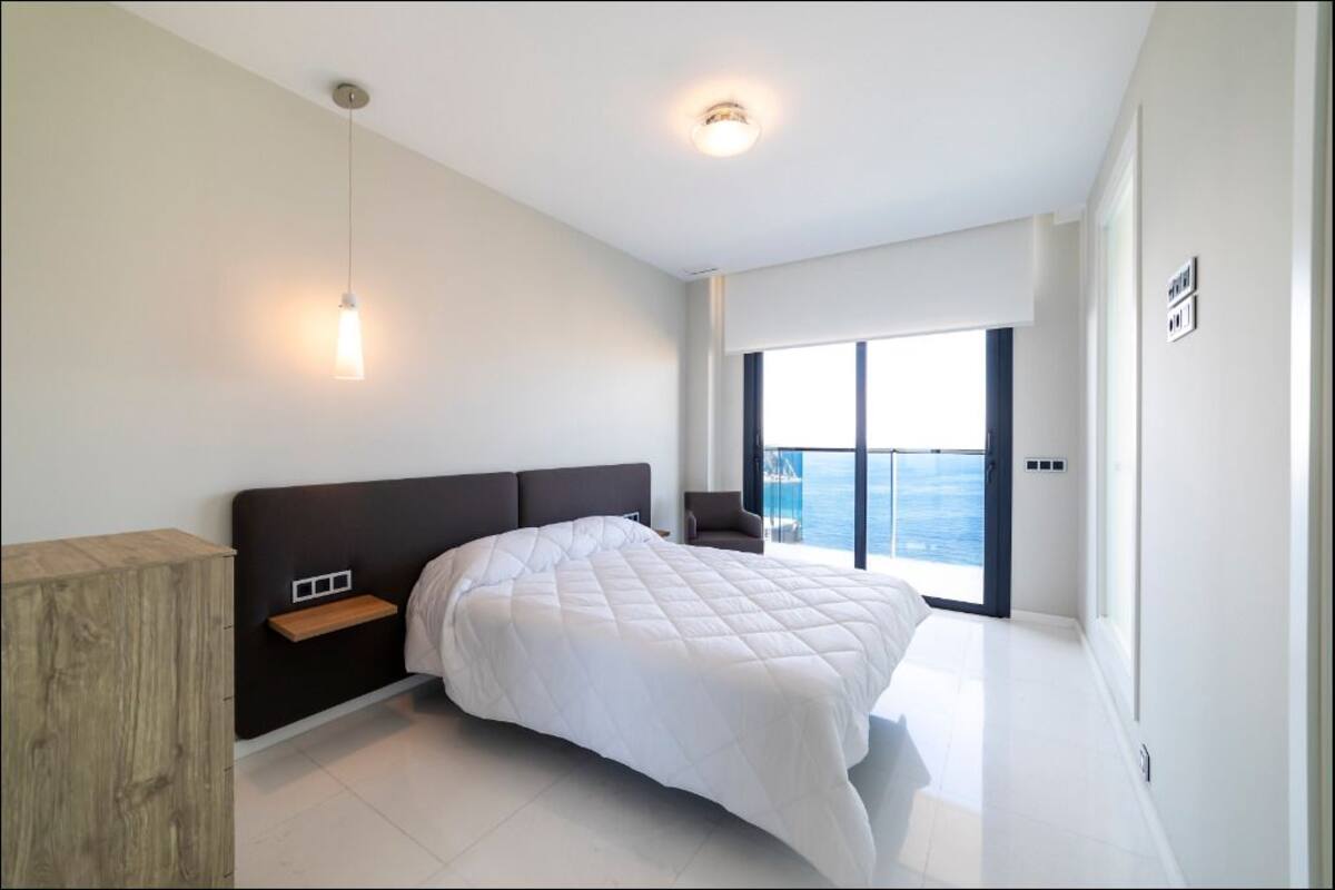 A modern bedroom with a large bed, a wooden nightstand, a hanging light, and a view of the sea through a balcony.