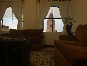 A living room with two brown sofas, a patterned rug, and a potted plant. A large window reveals a bell tower. - Amazing Lima - Near Plaza San Martin (Lima)
