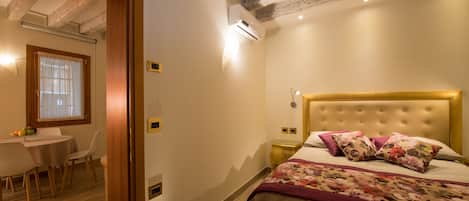 Un dormitori amb un llit, una petita taula de nit, una làmpada, una finestra amb cortines i una zona de menjador amb taula i cadires.