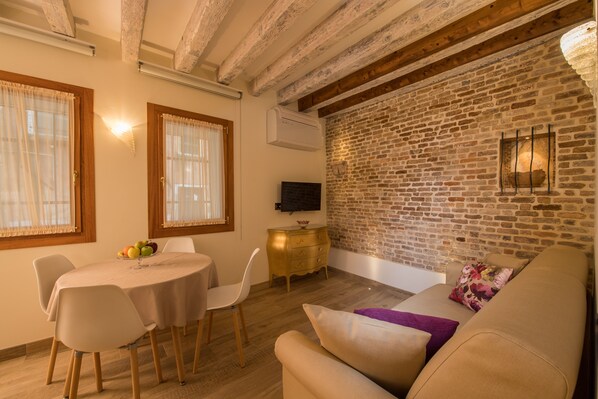 A cozy living room with a brick wall, a wooden ceiling, a round dining table with chairs, a sofa, and a TV mounted on the wall. - Nike Apartments Venice Castello (Venice)