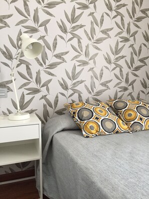 A bedroom with a patterned wallpaper, a bedside lamp, a grey bedspread, and patterned pillows.