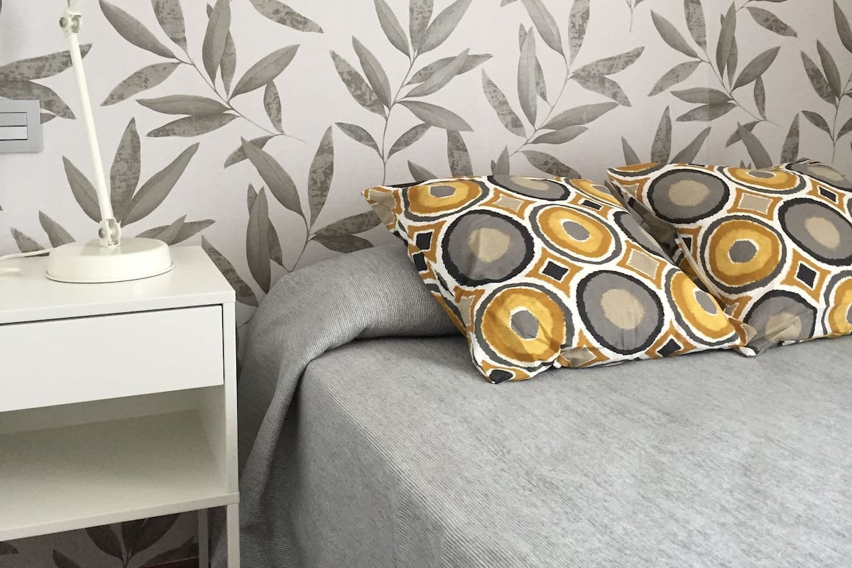 Makuuhuone, jossa on kuviollinen tapetti, yöpöytälamppu, harmaa päiväpeite ja kuviolliset koristetyynyt.