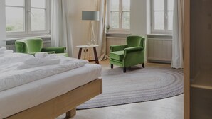 A hotel room with a bed, green armchairs, a standing lamp, and a view of the outdoors through large windows. - Die Aussicht (Neuburg)
