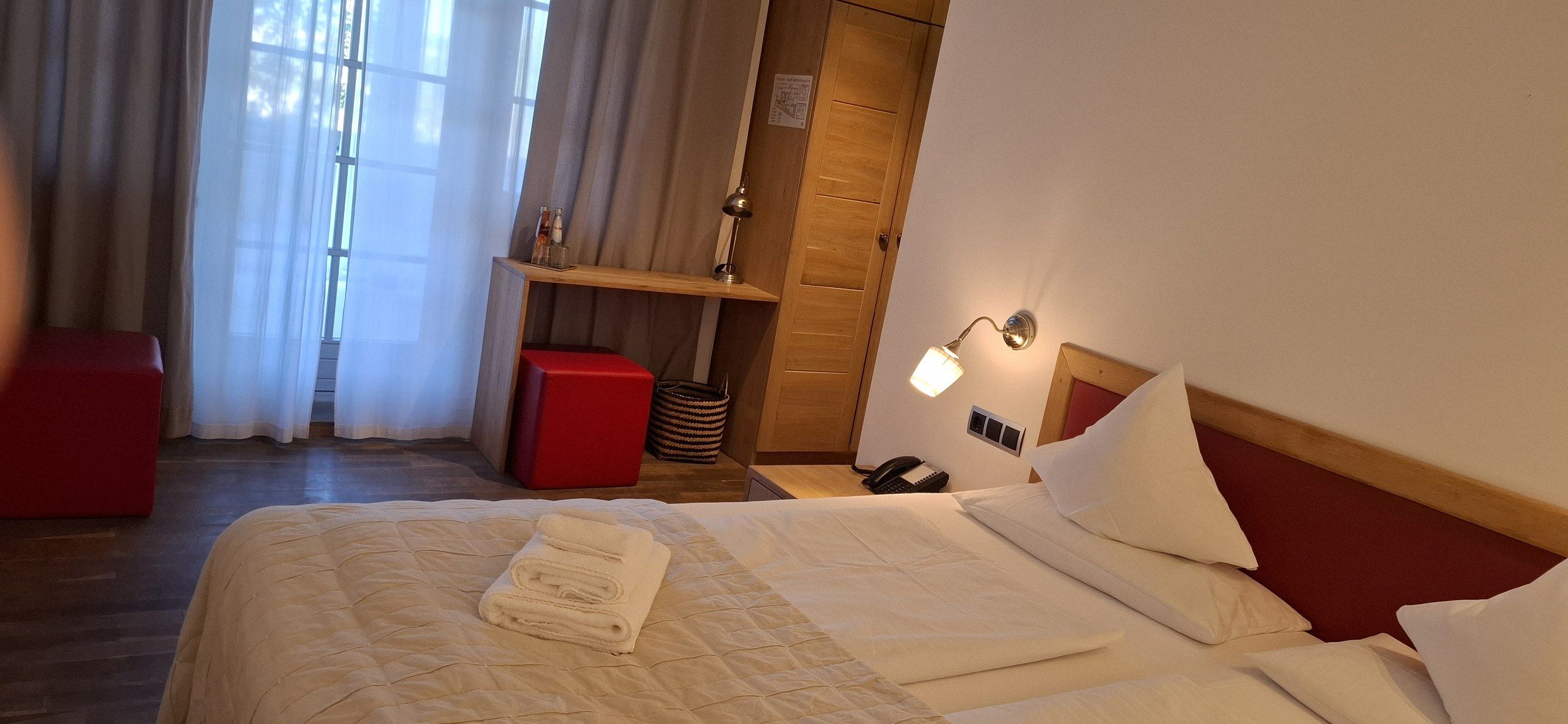 A hotel room with a bed, a red ottoman, a bedside lamp, and a wooden shelf with bottles.