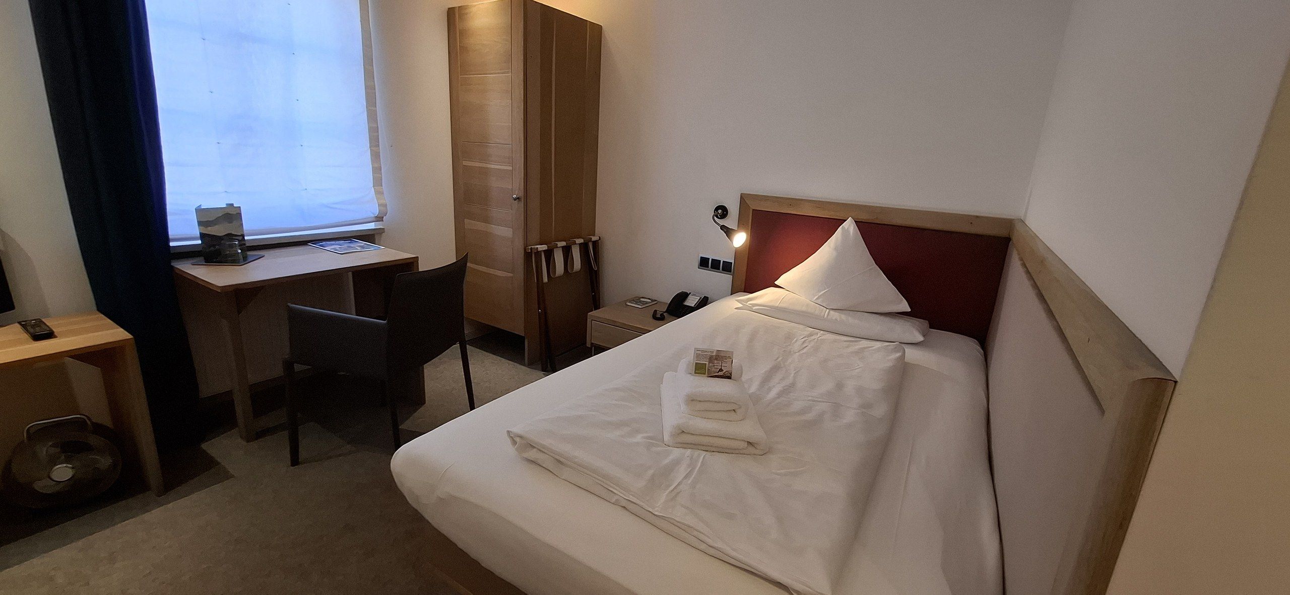 A hotel room with a bed, a desk, a chair, a wardrobe, and a bedside table.