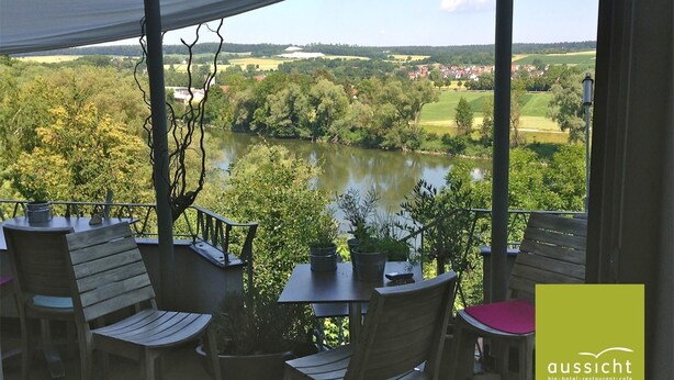 A terrace with wooden furniture, a table, and potted plants, overlooking a river and a rural landscape. - Die Aussicht (Neuburg)