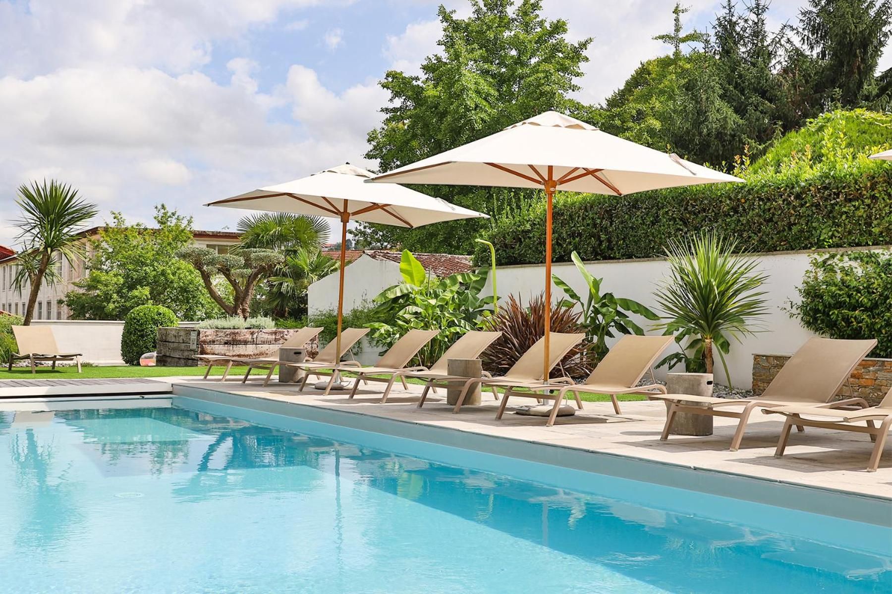 Una piscina con sillones de descanso y sombrillas, rodeada de follaje y una pared blanca.