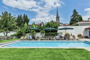 Une piscine entourée de chaises longues et de parasols, avec une végétation luxuriante et une église en arrière-plan.