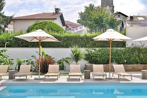 A poolside area with lounge chairs and umbrellas, surrounded by greenery and buildings. - Hôtel Argia (Hasparren)