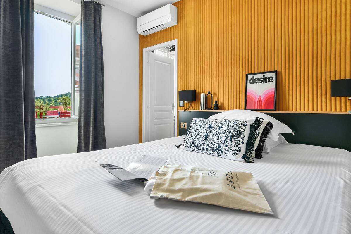 A hotel room with a bed, a window with curtains, a wall-mounted air conditioner, a framed picture, and a bedside table with a lamp and a phone.