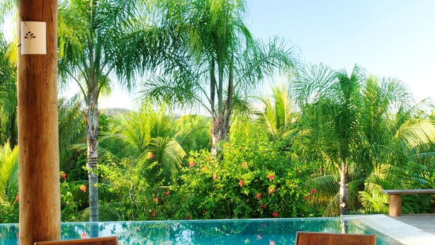 A wooden deck with two wooden chairs, a woven table, and a view of a swimming pool surrounded by lush greenery. - Txai Resort casa 10 (Itacaré)