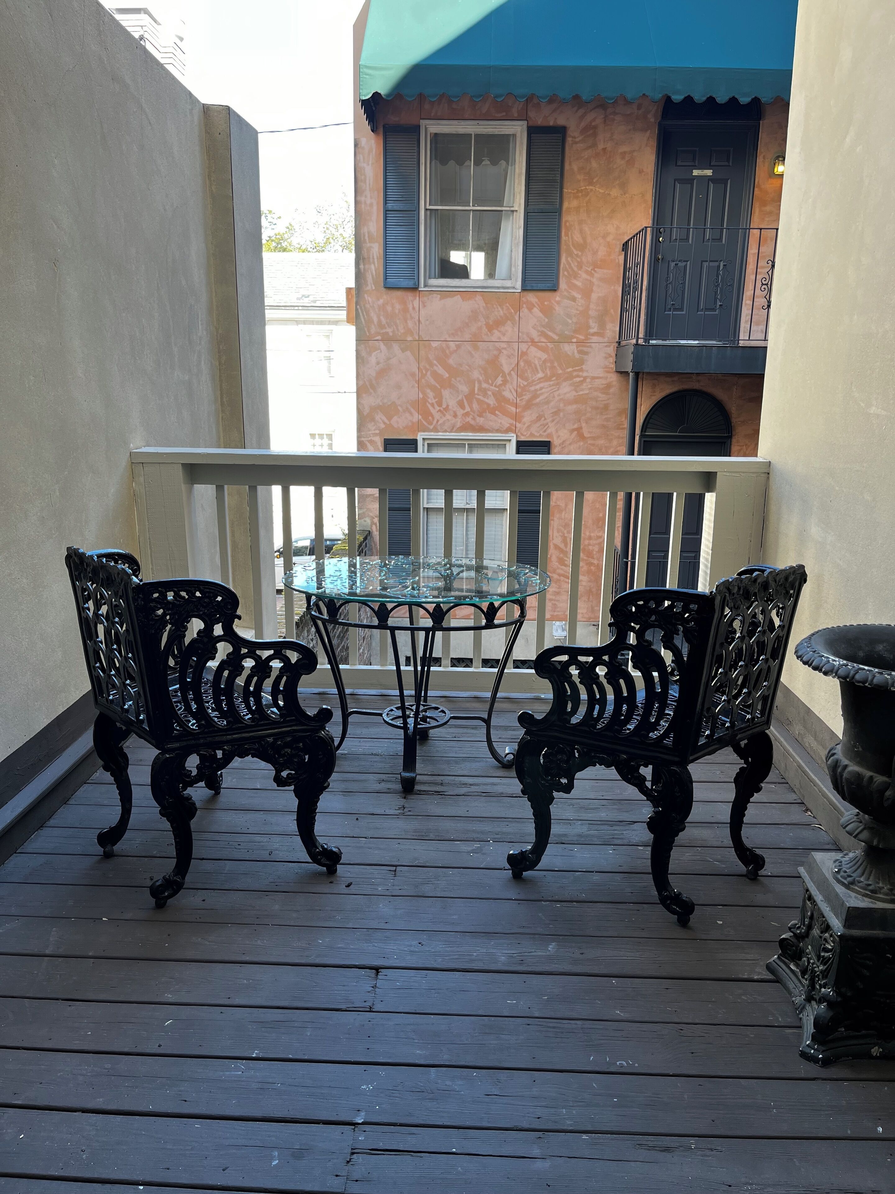 A balcony with two ornate metal chairs and a glass table, overlooking a building with a blue awning and a door.